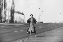 DDR Fotografie Bergemann Birgit Karbjinski Berlin 1984, schwarz-weiß Fotografie von einer Frau am Straßenrand mit den Händen in die Seite gestemmt, im Hintergrund steigt schwarzer Rauch aus einem Fabrikschornstein